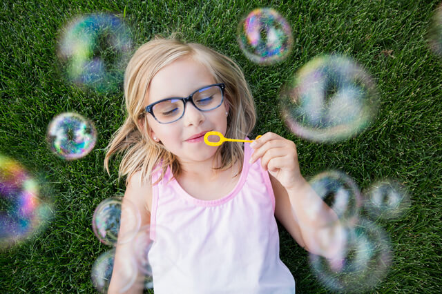 A young girl laying in grass, blowing bubbles from a wand.