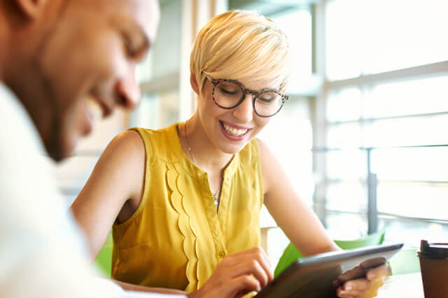 A young woman uses a tablet while a man smiles in the foreground.