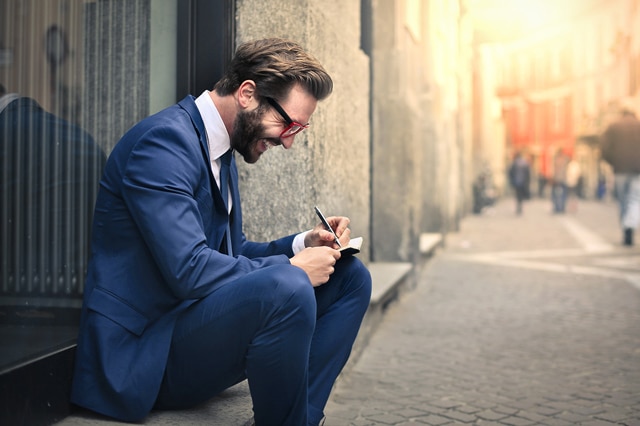 A young businessman wearing glasses sits outside, smiling and taking notes.