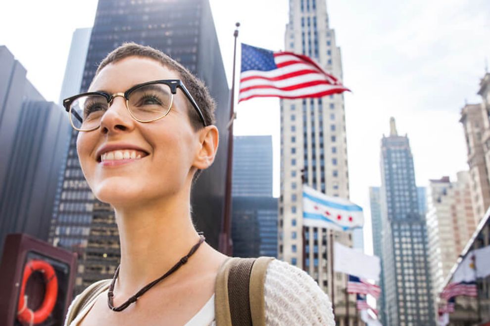 Young woman with glasses smiles in an outdoor city environment with flags in the background.