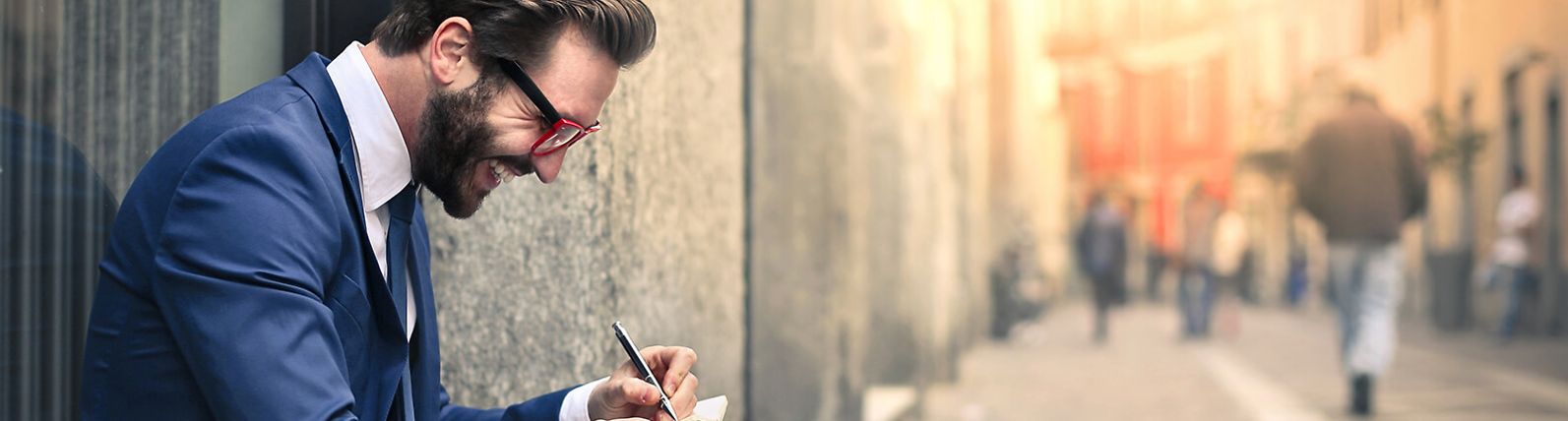 A young businessman wearing glasses sits outside, smiling and taking notes.