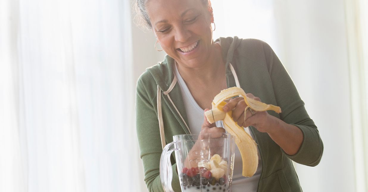 woman making fruit smoothie in blender and cutting up a banana